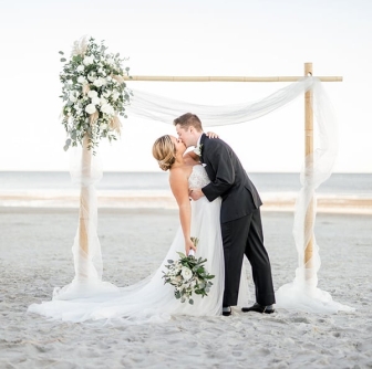 bride and groom kissing under a decorated arbor on the beach at The Dunes House