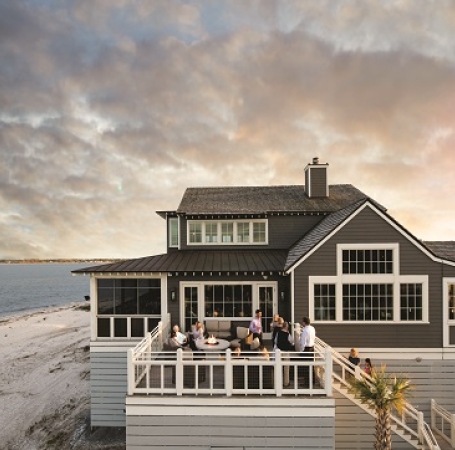 A big and luxury beach house with a group of people on the deck looking at the beach