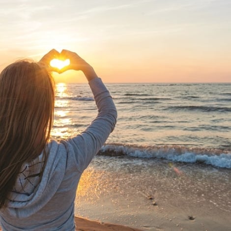 Girl standing on a beach making a hear shape with her hands facing the water