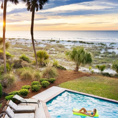 woman lounging on a pool floatie in a pool next to the beach during sunset