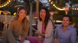 Three people sit under a string of lights while drinking out of wine glasses.