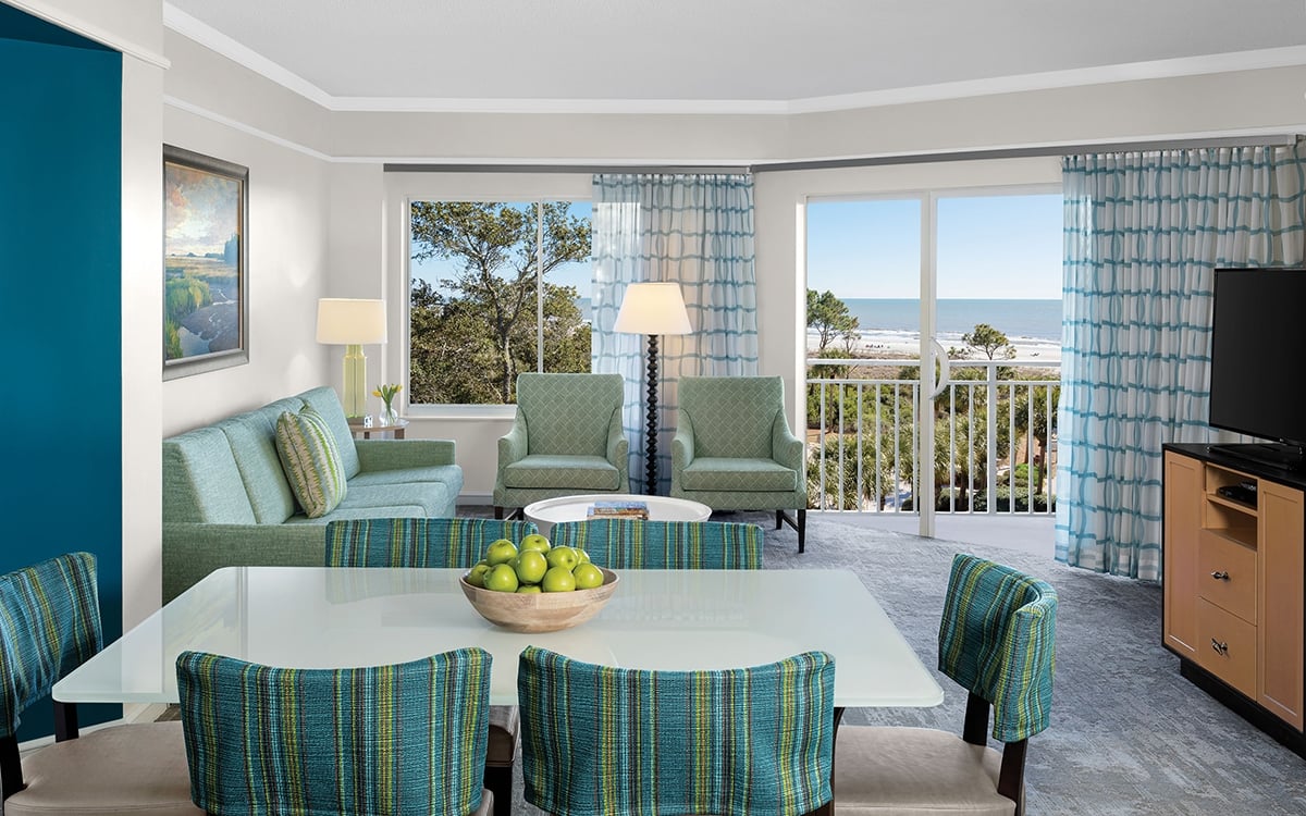 Villa living and dining area, stripped teal upholstered dining chairs, TV, sofa and two chairs, round cocktail table, teal blue and white curtains, balcony.