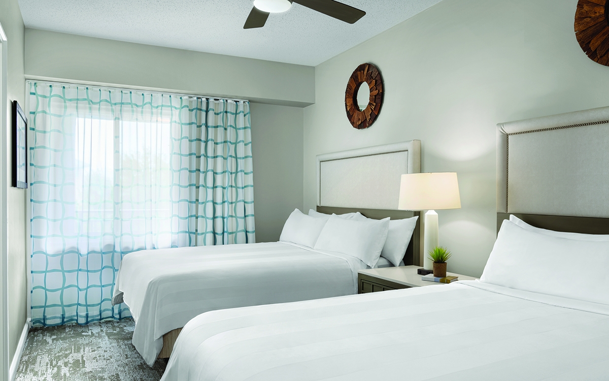 Guest room with double queen beds, dark wood round wall art, ceiling fan, teal blue and white curtains. 