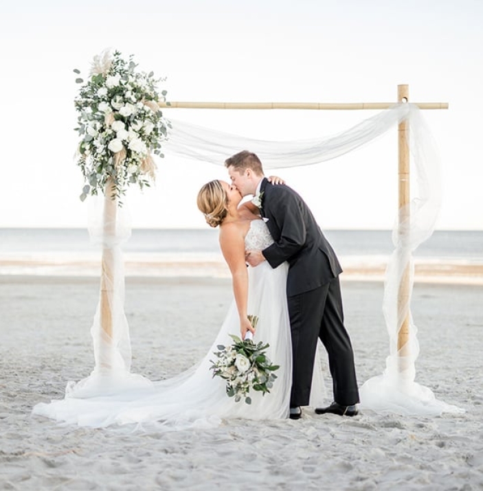 bride and groom kissing under a decorated arbor on the beach at The Dunes House